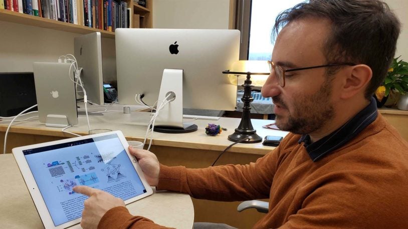 photo of Justin Kinney pointing to a model of an artificial neural network on his tablet