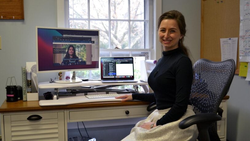 A photograph of Hannah Stewart, the Banbury Center's new Communications and Special Projects Coordinator. She is sitting in a black swivel chair and wearing a black turtleneck and cream skirt in front of a desk.
