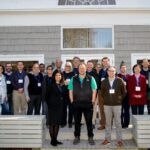 A photo of 26 people standing in three rows on the wood back patio of the Banbury Center's conference room. All meeting participants are wearing blue lanyards with name tags and are looking at the camera.