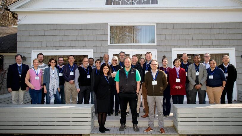 A photo of 26 people standing in three rows on the wood back patio of the Banbury Center's conference room. All meeting participants are wearing blue lanyards with name tags and are looking at the camera.