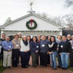 A photograph of 22 people standing in two rows in front of the Banbury Center. All meeting participants are wearing blue lanyards with name tags and are smiling.