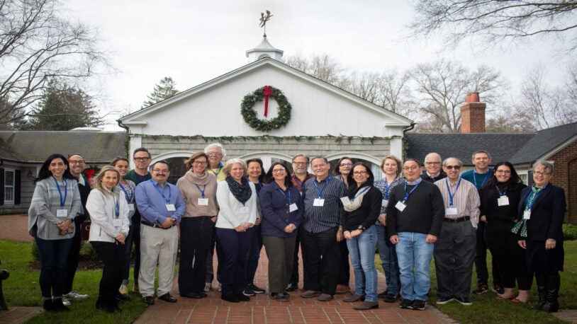 A photograph of 22 people standing in two rows in front of the Banbury Center. All meeting participants are wearing blue lanyards with name tags and are smiling.