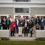 26 people standing in three rows on the wood back patio of the Banbury Center's conference room. All meeting participants are wearing blue lanyards with name tags and are smiling.