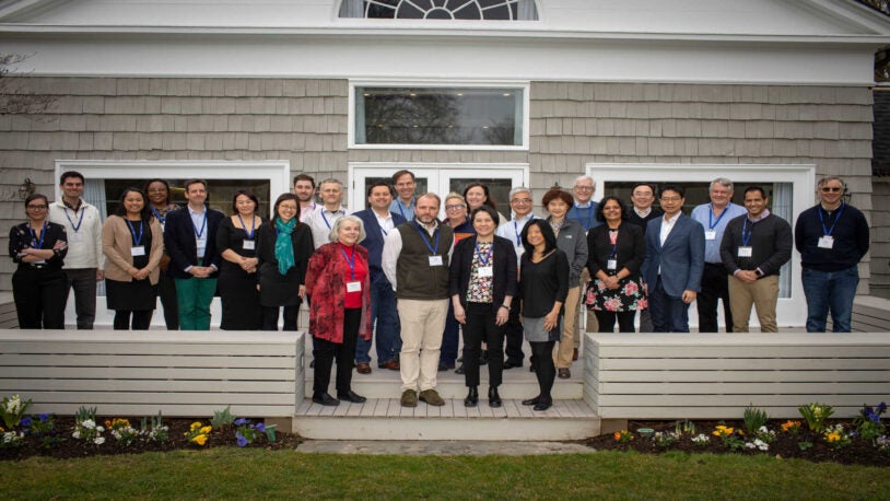 A photograph of 26 people standing in three rows on the wood back patio of the Banbury Center's conference room. All meeting participants are wearing blue lanyards with name tags and are smiling.
