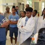 Assistant Director Jason Williams demonstrates a scientific object to attentive students in a lab, some wearing lab coats.