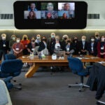 A photograph of 22 people standing in two rows in front of the Banbury Center. All meeting participants are wearing masks and blue lanyards with name tags. There are seven people displayed on the projector, smiling.