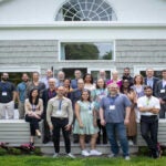 23 people standing in three rows on the wood back patio of the Banbury Center's conference room. All meeting participants are wearing blue lanyards with name tags and are smiling.