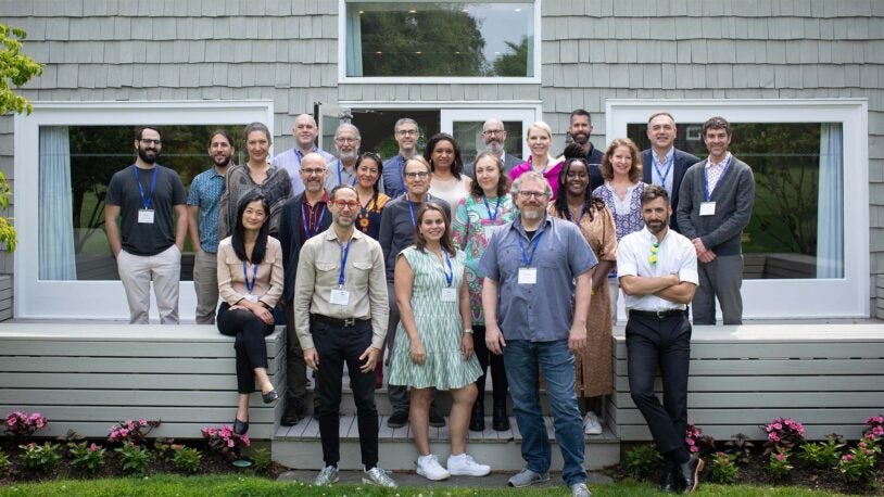 A photograph of 23 people standing in three rows on the wood back patio of the Banbury Center's conference room. All meeting participants are wearing blue lanyards with name tags and are smiling.