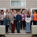 31 people standing in three rows on the wood back patio of the Banbury Center's conference room. All meeting participants are wearing blue lanyards with name tags and are smiling.