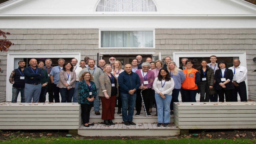 A photograph of 31 people standing in three rows on the wood back patio of the Banbury Center's conference room. All meeting participants are wearing blue lanyards with name tags and are smiling.