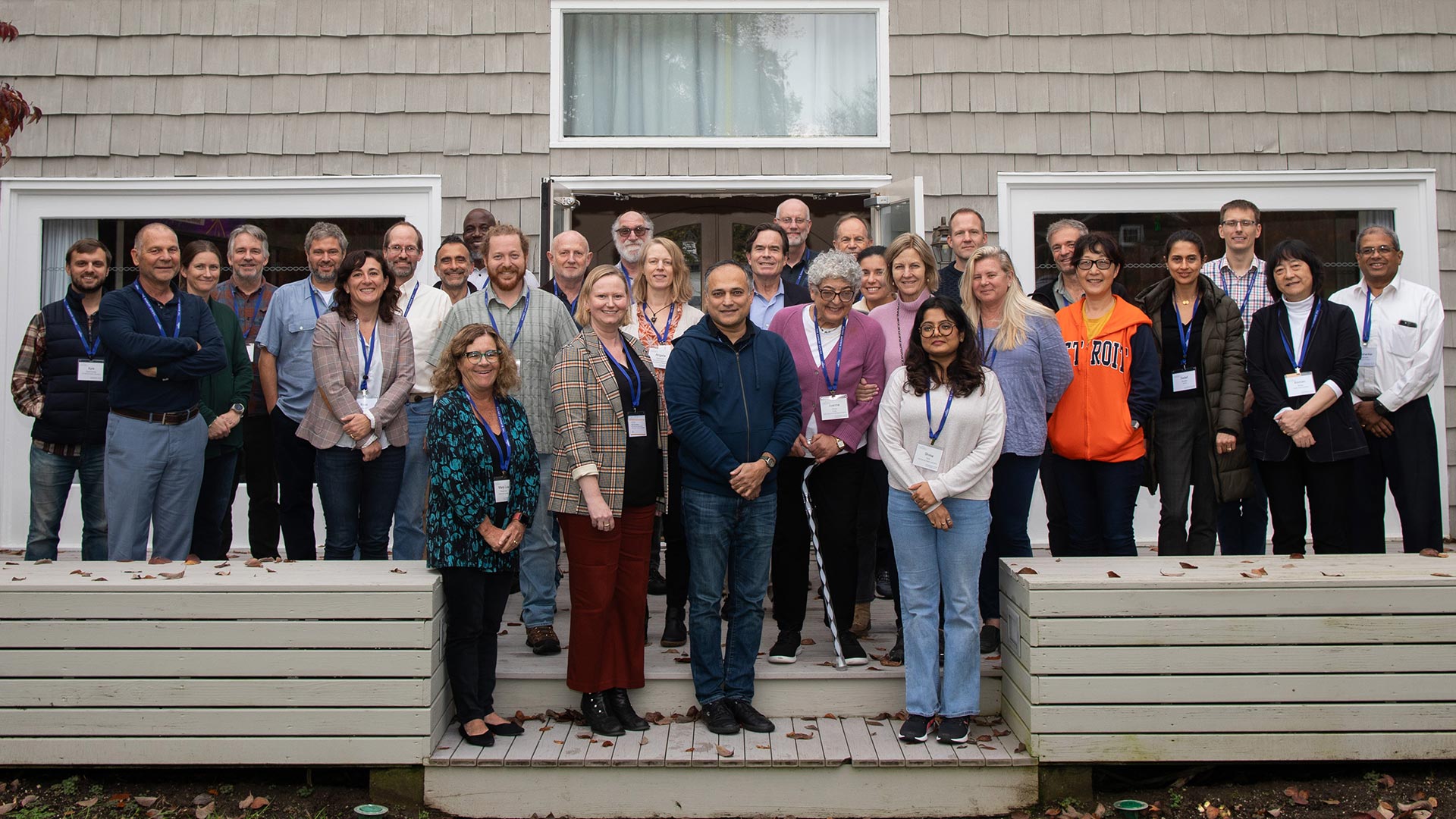 A photograph of 31 people standing in three rows on the wood back patio of the Banbury Center's conference room. All meeting participants are wearing blue lanyards with name tags and are smiling.