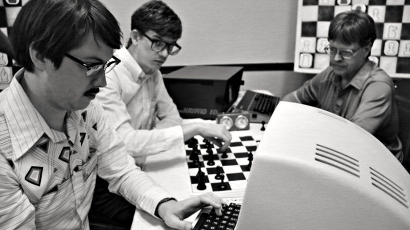 film still from Computer Chess film, 3 men sitting with chess boards and 1980s computers