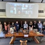 13 people standing in one row in the conference room. All are wearing masks and lanyards. Behind them, there are 14 people on the projector participating in the meeting virtually.