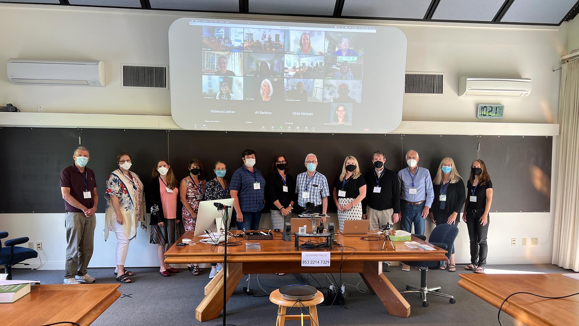 13 people standing in one row in the conference room. All are wearing masks and lanyards. Behind them, there are 14 people on the projector participating in the meeting virtually.