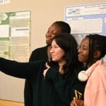 Three high school students stand together smiling as they take a selfie in front of DNALC scientific research posters displayed on a wall behind them. One student gives a thumbs-up gesture while another wears headphones around their neck.