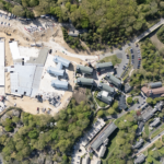 Aerial view of a construction site next to completed buildings surrounded by trees and a lake.