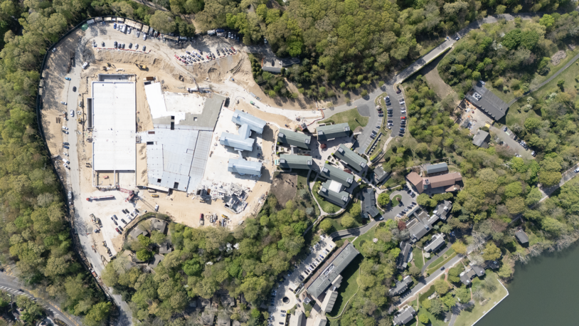 Aerial view of a construction site next to completed buildings surrounded by trees and a lake.
