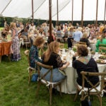 Large group of people sit at round tables under a tent at an outdoor garden event or luncheon.