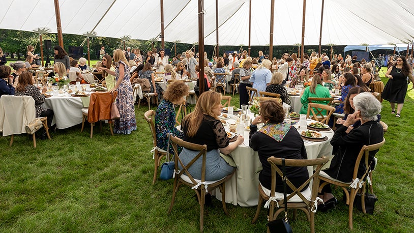 Large group of people sit at round tables under a tent at an outdoor garden event or luncheon.