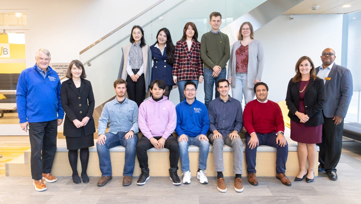 A group of thirteen people pose together indoors on steps by a glass railing, smiling at the camera.