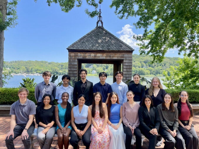 A group of 17 CSHL Undergraduate Research Program (URP) participants, Summer 2025 pose together outdoors in front of a wooden gazebo and the harbor.