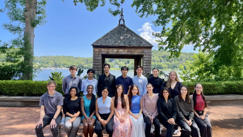 A group of 17 young adults pose outdoors in front of a wooden structure, trees, and a lake on a sunny day.