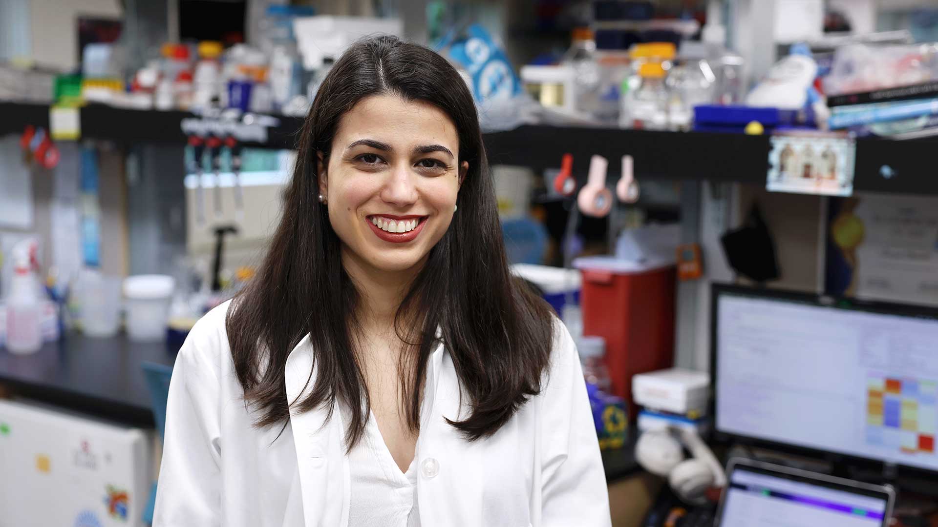 CSHL Assistant Professor Corina Amor Vegas in a lab coat smiles in a laboratory with equipment, bottles, and a computer in the background.