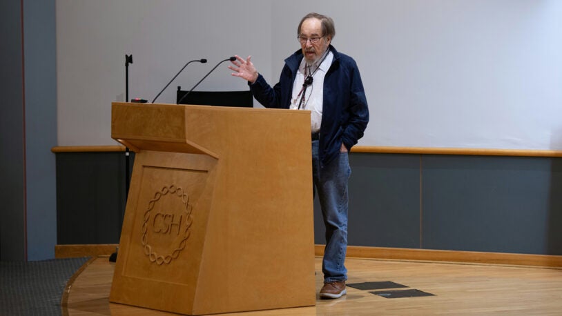 David Baltimore speaks at a wooden podium in an auditorium with a microphone and blank screen behind him.