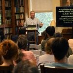 Marc Torres speaks to an audience in a library; a screen displays a book titled Long Island Eugenics.