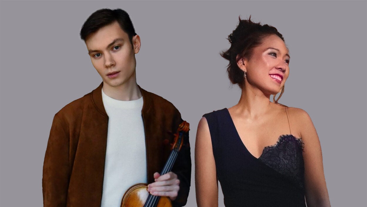 A man holding a violin stands next to a smiling woman in a black dress, both on a gray background.