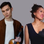 A man holding a violin stands next to a smiling woman in a black dress, both on a gray background.