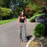 Kristin Smith in black top and floral pants stands by a mailbox on a sunny, tree-lined suburban street.