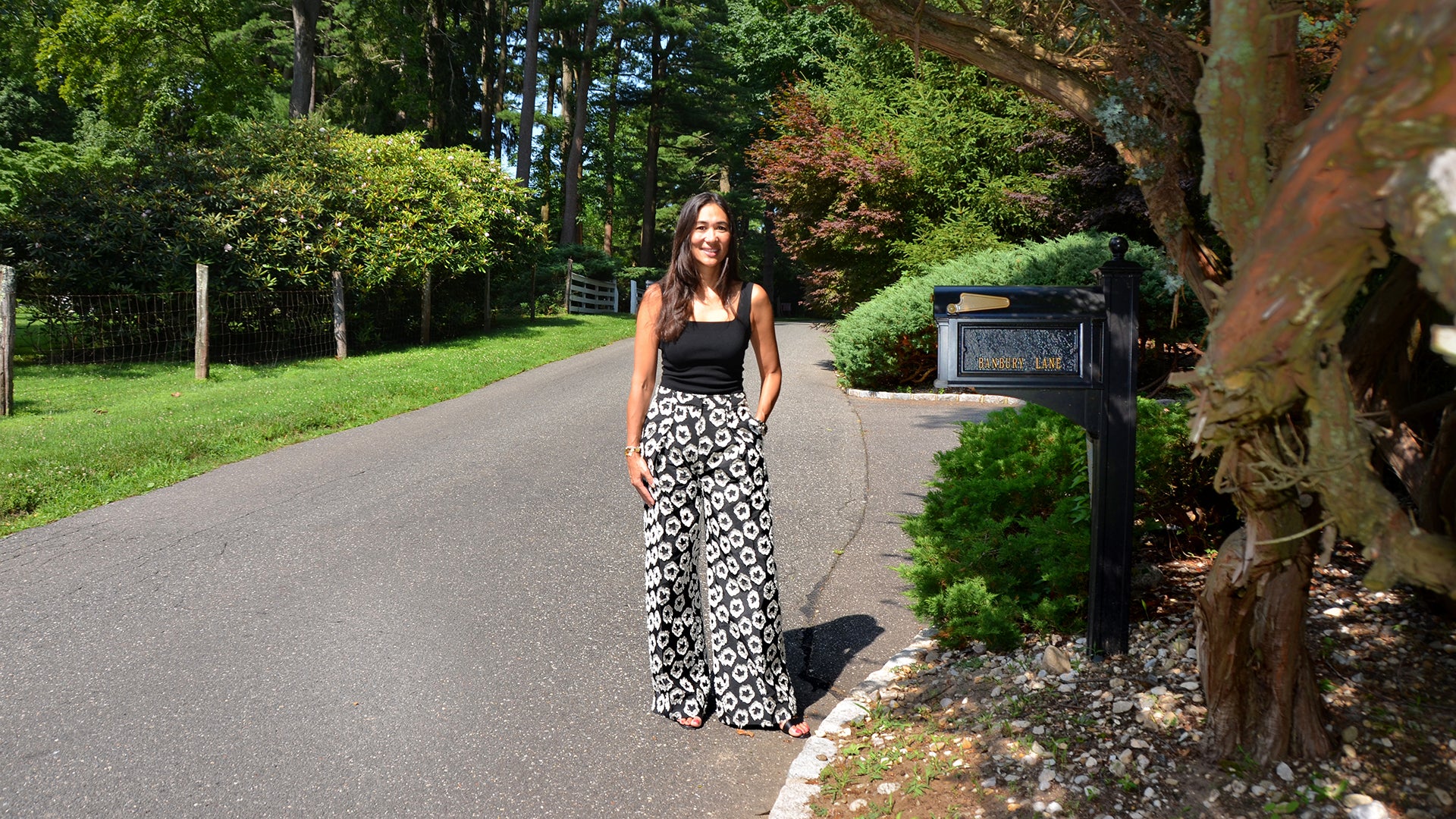 Kristin Smith in black top and floral pants stands by a mailbox on a sunny, tree-lined suburban street.