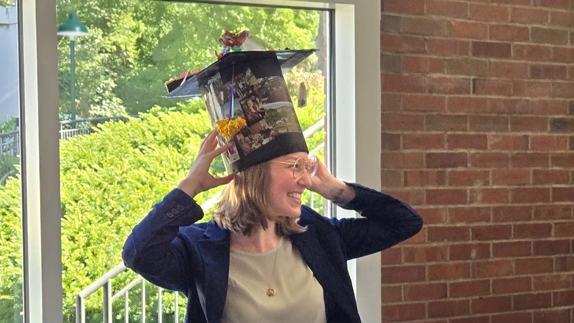 Leah Braviner indoors wearing a decorated graduation cap, hands touching the sides, sunlight in the background.