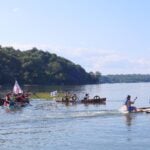 People race homemade rafts on a harbor near a wooded shoreline under a clear blue sky.