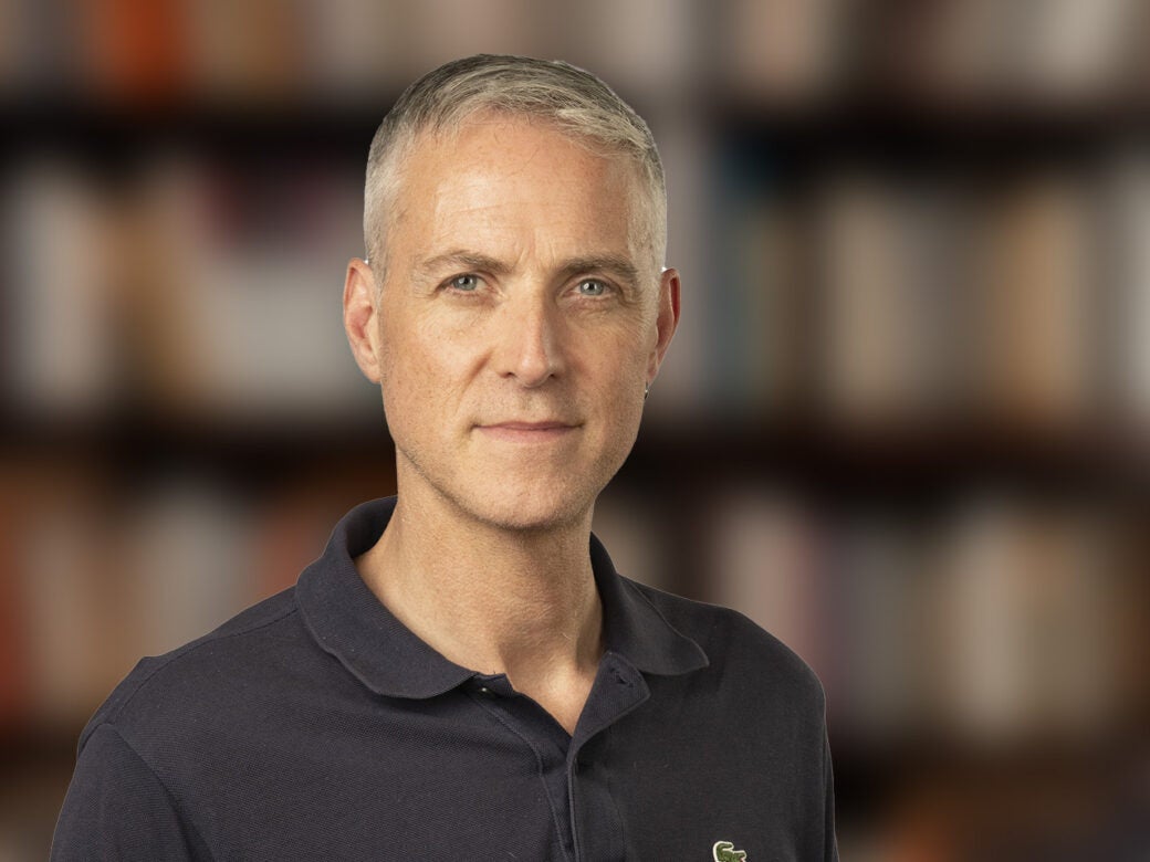 A middle-aged man with short gray hair wearing a dark polo shirt stands in front of blurred bookshelves.