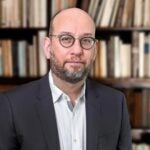 David Crotty, a man in glasses and a suit stands in front of a blurred bookshelf filled with books.