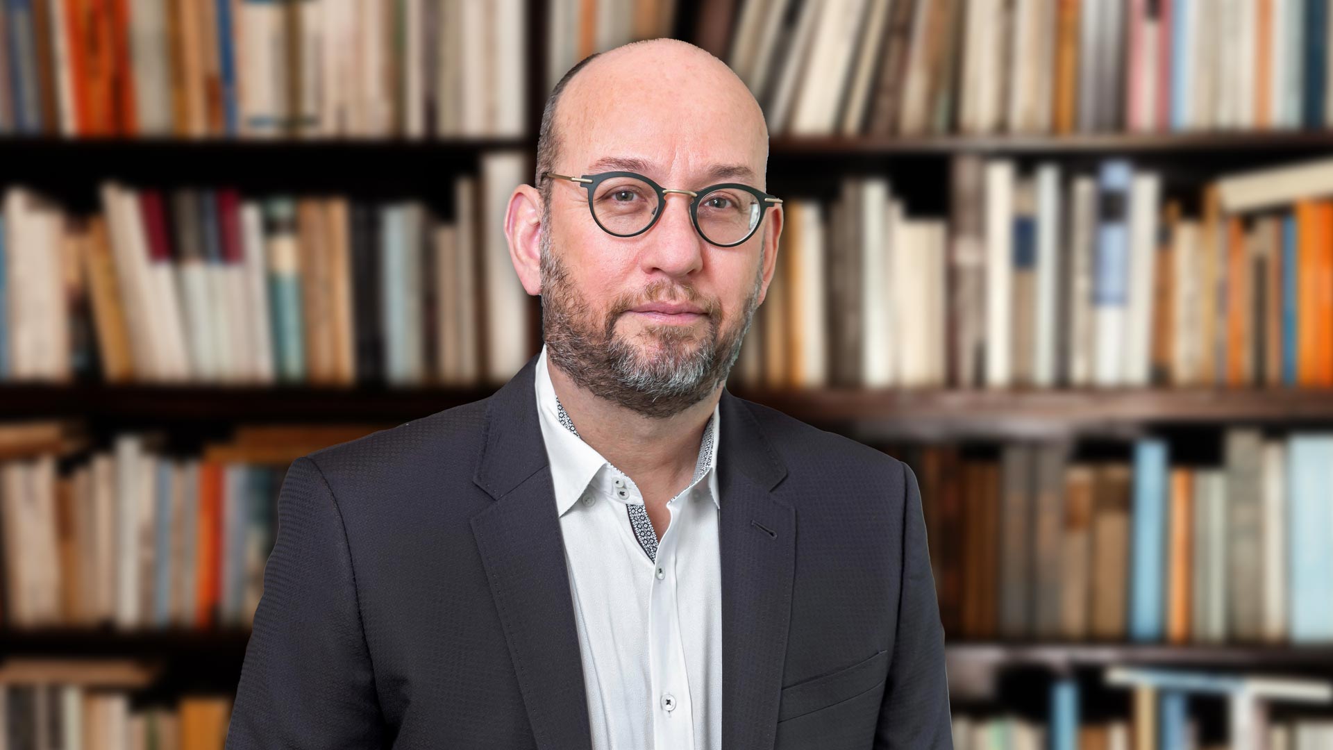 David Crotty, a man in glasses and a suit stands in front of a blurred bookshelf filled with books.