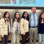 Seven people, including five young women and two men, stand smiling together in front of a screen.