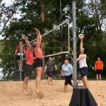 People play beach volleyball outdoors; one player jumps to spike the ball as others attempt to block.