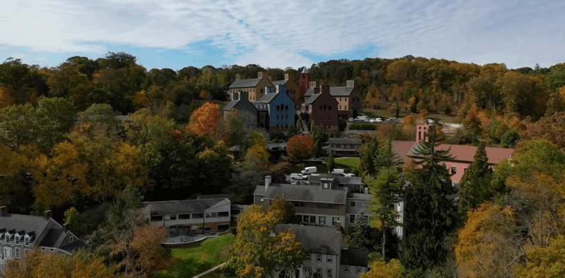 Aerial view of colorful buildings and houses surrounded by autumn trees and rolling hills under a partly cloudy sky.