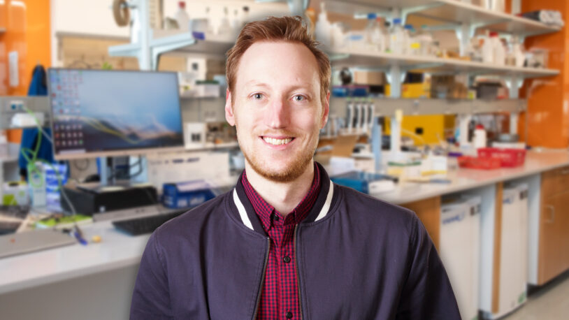 Jeremy Borniger with light hair stands in a bright laboratory with shelves, equipment, and computers in the background.