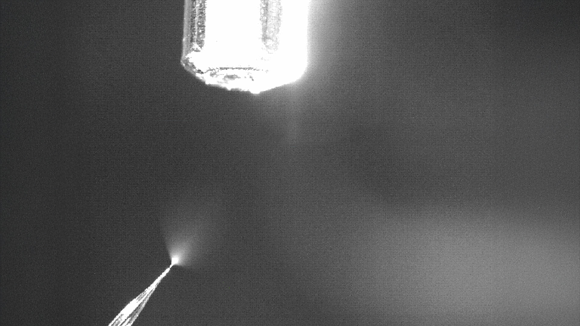 A bright nozzle sprays a fine mist or liquid downward in a dim, monochrome setting.