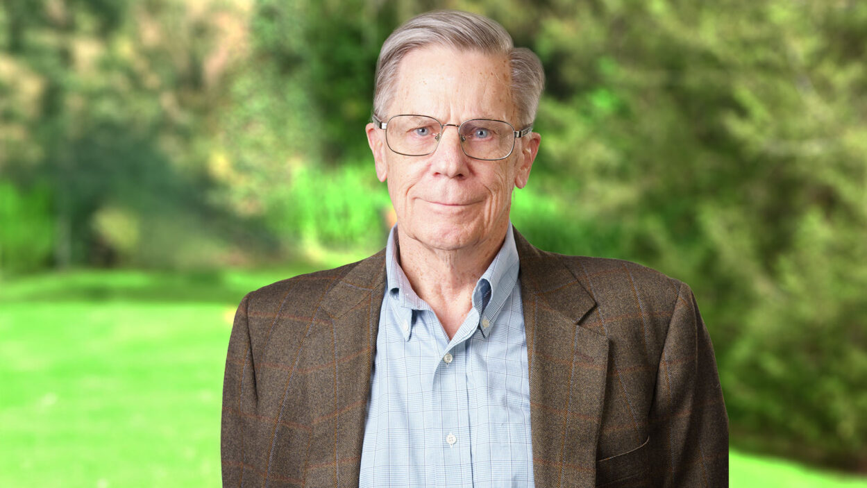 An older man with glasses stands outdoors, wearing a plaid blazer and collared shirt, with greenery behind him.
