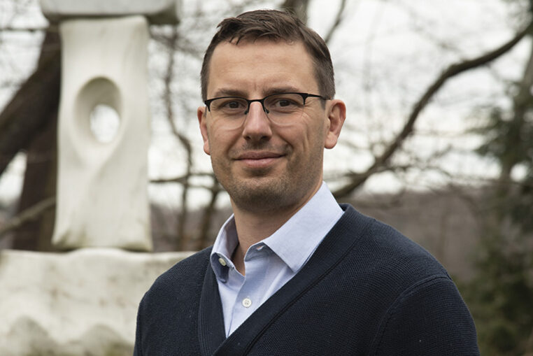 Paolo Cifani with glasses and short hair wearing a blue sweater and collared shirt, standing outdoors near a sculpture.