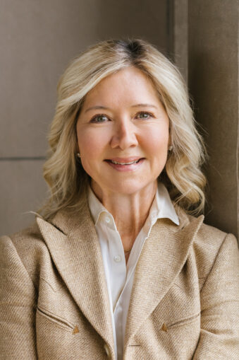 Terri Keogh with blonde hair in a beige blazer and white shirt, standing indoors by a wall.