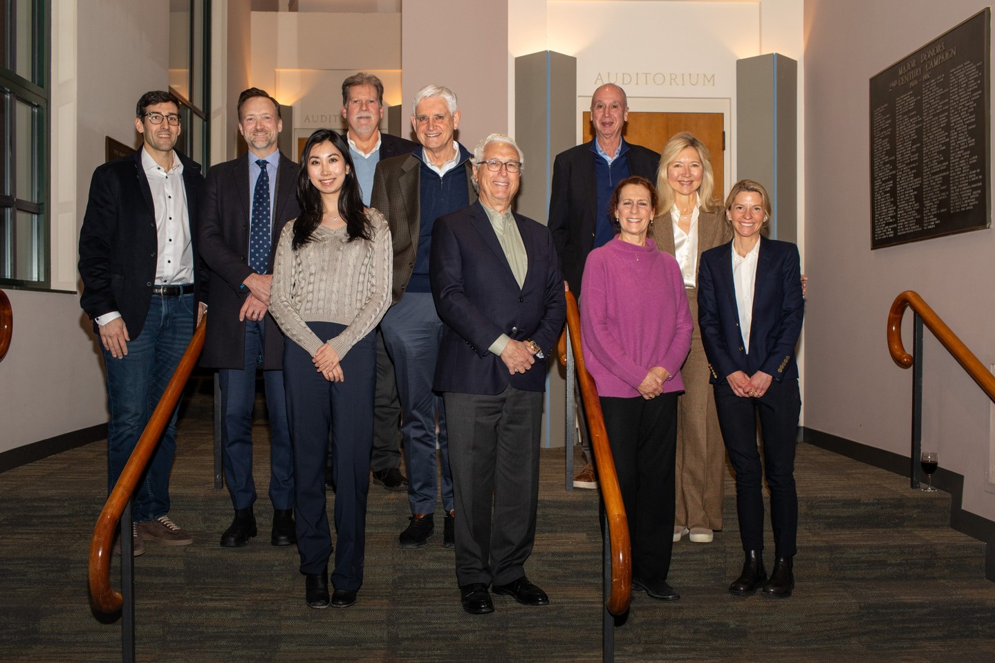 Ten people stand in two rows on an indoor staircase in front of an auditorium entrance.