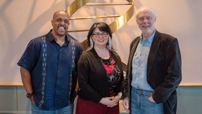 Jason Williams, Charla Lambert, and John Inglis stand together indoors in front of a decorative gold sculpture on the wall.