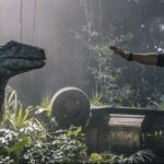 A man faces a velociraptor in a jungle with fallen park equipment and misty light in the background.