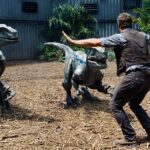 A man faces three velociraptors with his arms raised in a fenced, tropical enclosure.